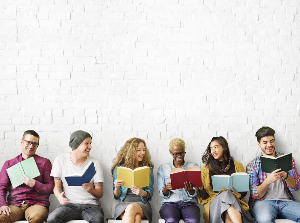 A book club of men and woman hanging out chatting and laughing in front of a white wall